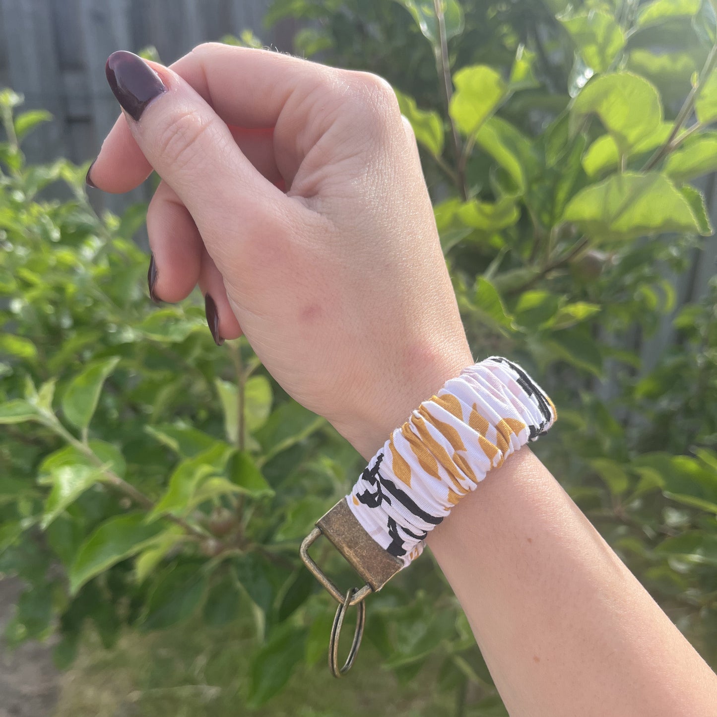 Sleutelhanger in de vorm van een Scrunchie voor om je pols. Van stof met elastiek. Wit met bladeren patroon in okergeel, pastel roze en zwart. Met bronskleurige klem/ring. Handgemaakt door Studio Dwarsdenker.