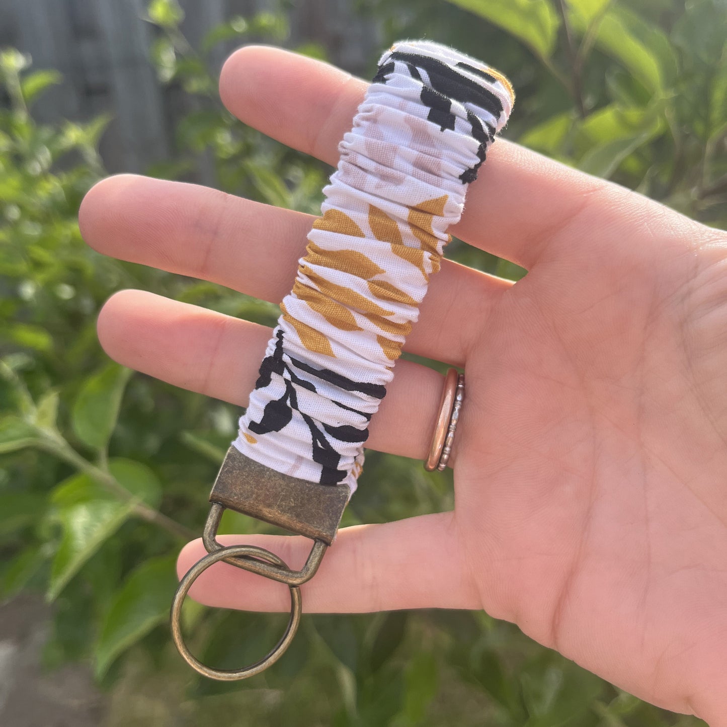 Sleutelhanger in de vorm van een Scrunchie voor om je pols. Van stof met elastiek. Wit met bladeren patroon in okergeel, pastel roze en zwart. Met bronskleurige klem/ring. Handgemaakt door Studio Dwarsdenker.