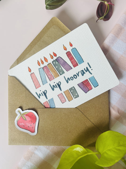 Ansichtkaart met een witte achtergrond met daarop een kleurrijke waterverf illustratie van verjaardagskaarsjes en de tekst: hip hip hooray. Ontwerp door Studio Dwarsdenker