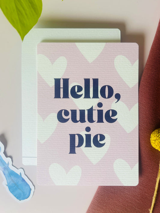 Ansichtkaart in zacht roze tint, met witte harten en de tekst: Hello, cutie pie. Ontwerp door Studio Dwarsdenker