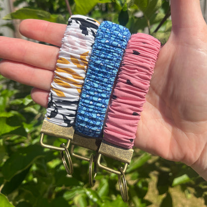 Sleutelhangers in de vorm van een  Scrunchie voor om je pols. Van stof met elastiek in cobalt blauw., roze met hartjes en wit met bladeren patroon in geel, pastel roze en zwart. Met bronskleurige klem/ring. Handgemaakt door Studio Dwarsdenker.