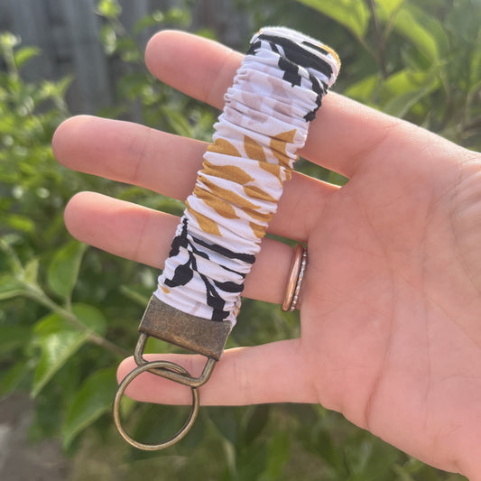 Sleutelhanger in de vorm van een  Scrunchie voor om je pols. Van stof met elastiek. Wit met bladeren patroon in okergeel, pastel roze en zwart. Met bronskleurige klem/ring. Handgemaakt door Studio Dwarsdenker.