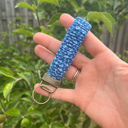 Sleutelhanger in de vorm van een  Scrunchie voor om je pols. Van stof met elastiek in blauwtinten met schubbenpatroon. Met bronskleurige klem/ring. Handgemaakt door Studio Dwarsdenker.