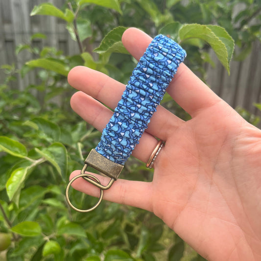 Sleutelhanger in de vorm van een  Scrunchie voor om je pols. Van stof met elastiek in blauwtinten met schubbenpatroon. Met bronskleurige klem/ring. Handgemaakt door Studio Dwarsdenker.